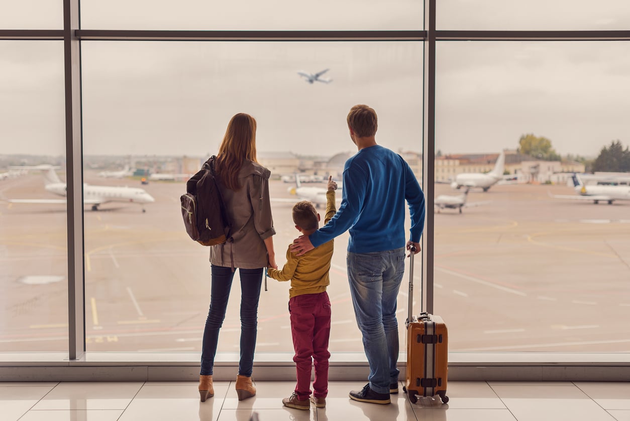 A family of three—a woman, man, and child—stand at an airport window watching planes on the runway. With their journey possibly affected by delayed purchases, the man has a suitcase, the woman carries a backpack, and the child holds their hands.