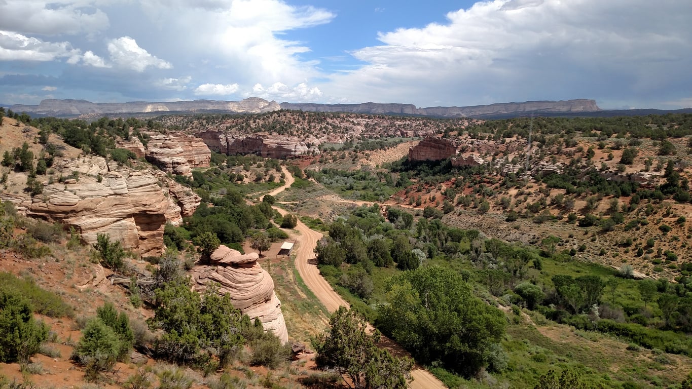 A winding dirt road curves through a rugged desert canyon with layered rock formations, scattered green trees and bushes, under a partly cloudy blue sky. Mountains are visible in the distance.