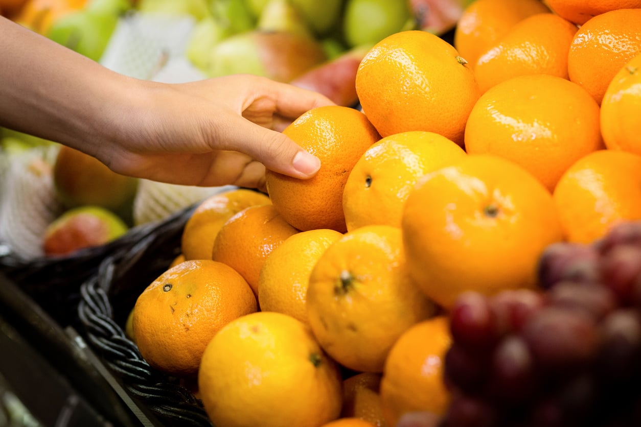 A person’s hand selecting a shiny orange from a pile of fresh oranges at a grocery store, with other fruits visible in the background.