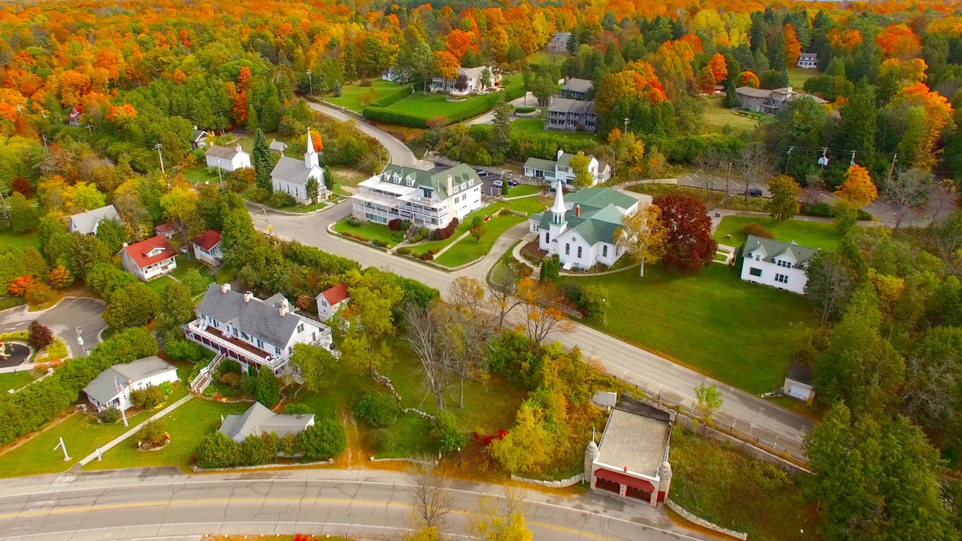 Aerial view of a small town with houses, churches, and roads surrounded by trees with autumn foliage in vibrant orange, red, and green colors. The area has lush green lawns and winding streets through the neighborhood.