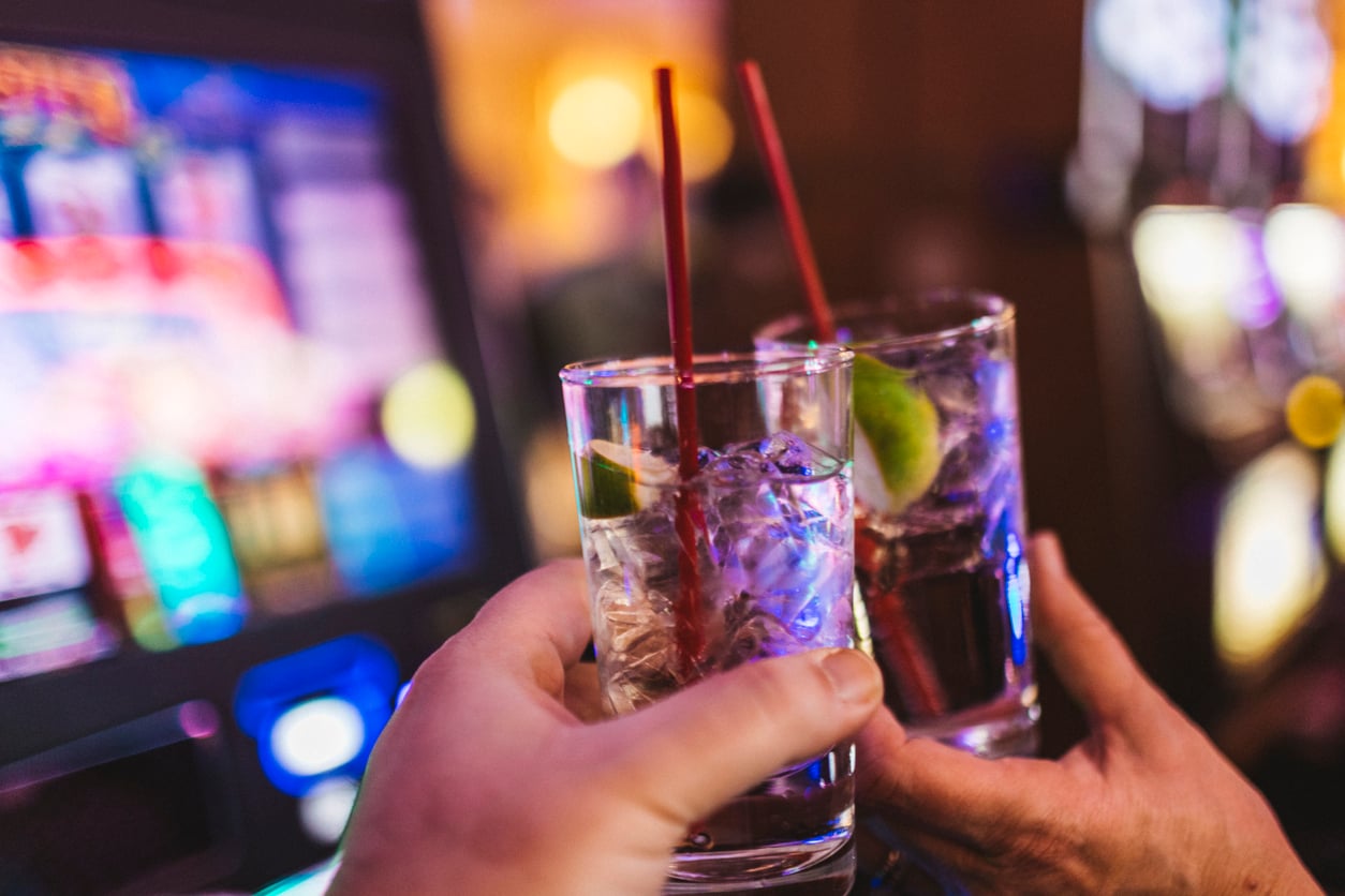 Two hands raise glasses with clear cocktails, ice, lime wedges, and red straws in front of a glowing slot machine—a lively scene that captures the thrill of enjoying Vegas nightlife while keeping Las Vegas budget tips in mind.