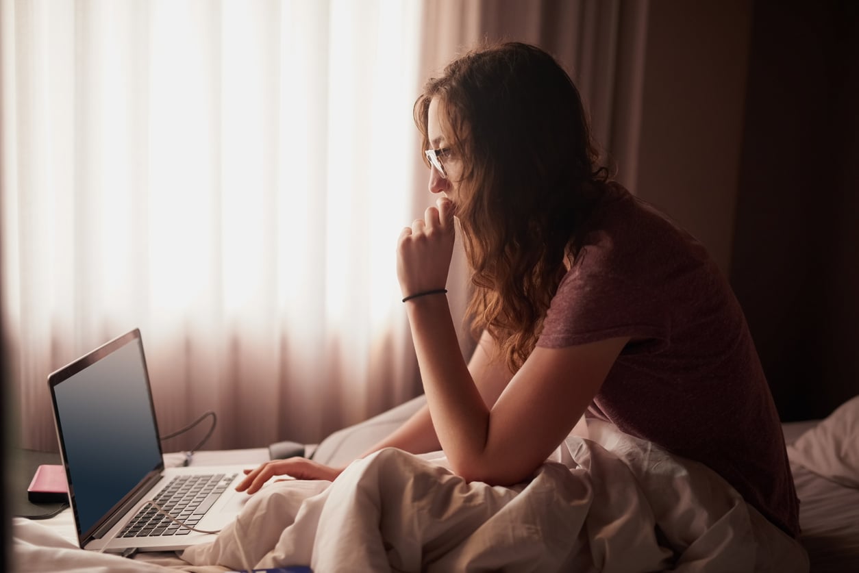 A woman with long hair and glasses sits on a bed covered with a white blanket, looking thoughtfully at a laptop—perhaps searching for concert ticket hacks. Soft daylight filters through curtains, creating a calm, cozy atmosphere.