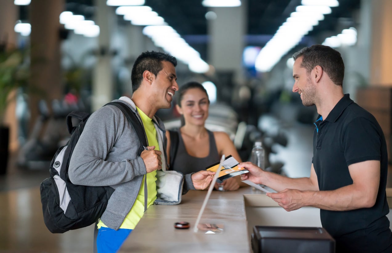 Two people at a gym reception desk smile as they talk with a staff member. One holds an ID card and pen—classic products men still buy—while the other, with a water bottle, stands behind. Gym equipment is blurred in the background.