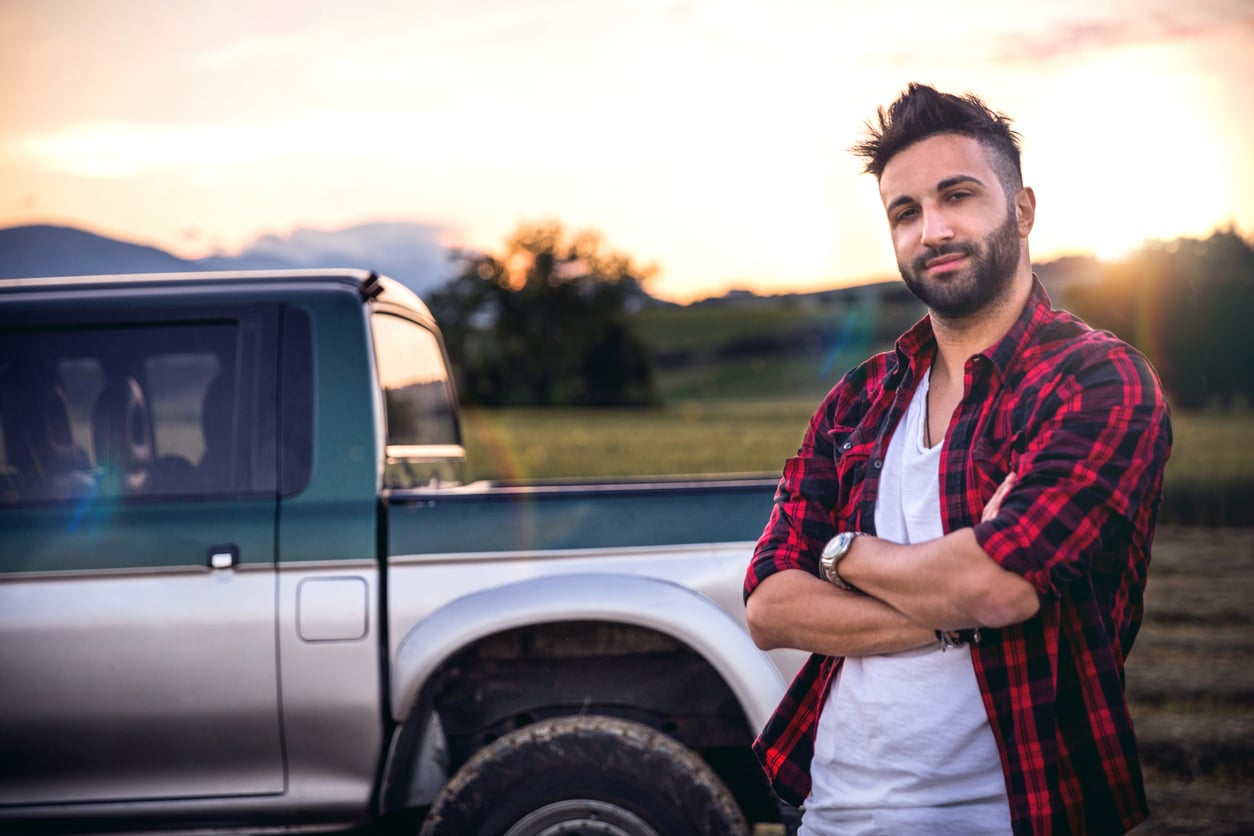 A man with dark hair and a beard, wearing a red plaid shirt over a white t-shirt, stands with arms crossed in front of a pickup truck—an image that reflects classic products men still buy—in a rural field at sunset.
