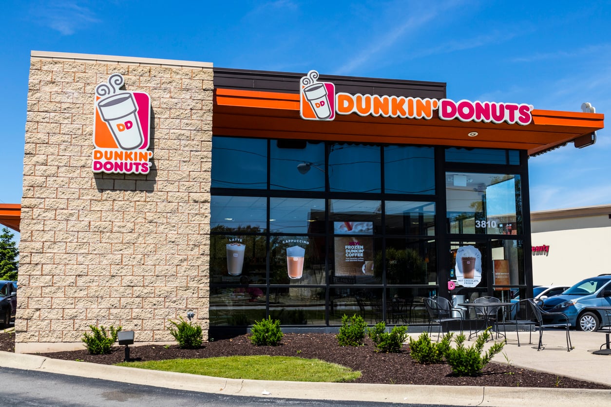 A Dunkin’ Donuts store with large glass windows, tan brick walls, and Dunkin’ Donuts signs above the entrance. Cars are parked nearby and the sky is clear and blue.