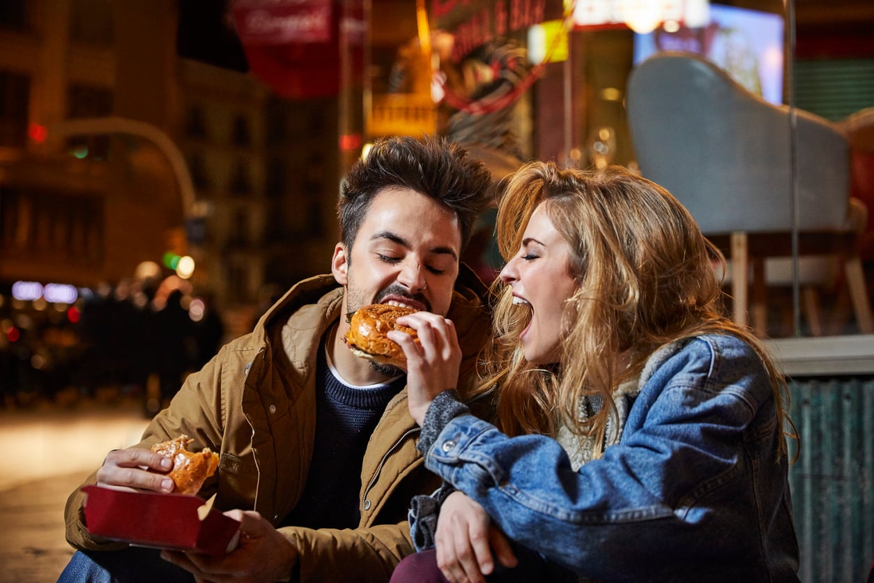A young man and woman sit close together outdoors at night, laughing and smiling as they enjoy burgers at 1991 prices, sharing a joyful moment in a lively, urban setting.