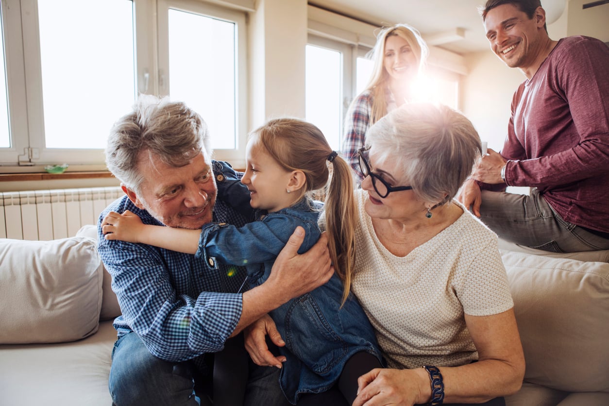 American family life. A young girl hugs her smiling grandfather on a couch while her grandmother sits beside them. Two adults stand in the background, all appearing happy in a bright, cozy living room.