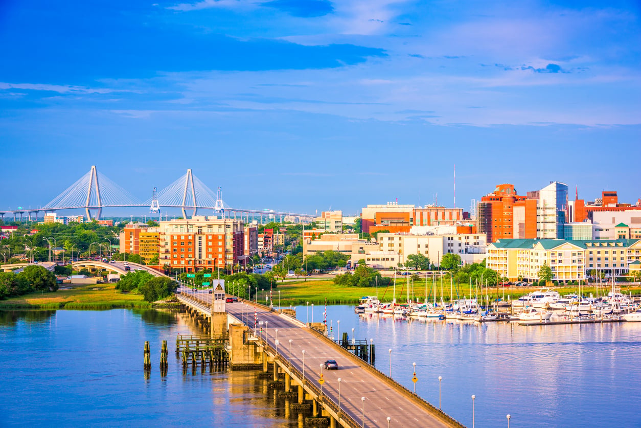 View of a city waterfront with a bridge extending over the water, boats docked at a marina, modern buildings in the background, and a cable-stayed bridge glistening under Florida Alternatives’ bright blue sky.