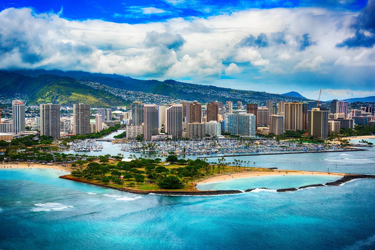 Aerial view of Honolulu, Hawaii—one of the top Small Business States—showing a skyline of tall buildings, a marina, sandy beaches, turquoise ocean waters, and lush green mountains under a partly cloudy sky.
