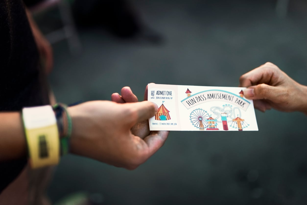 Close-up of two hands exchanging a colorful FunPass Amusement Park ticket, featuring images of a circus tent, Ferris wheel, and carousel; one hand wears wristbands—a perfect moment for sharing theme park hacks.