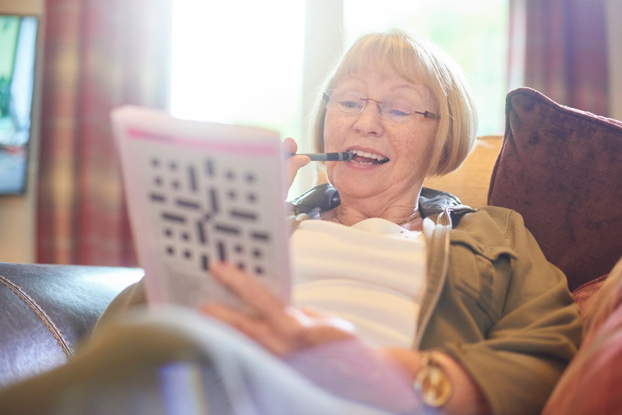 An older woman sits on a couch, smiling as she works on a crossword puzzle in a book—an enjoyable way to support mental health. She holds a pen to her mouth, appearing relaxed with natural light streaming through the window behind her.
