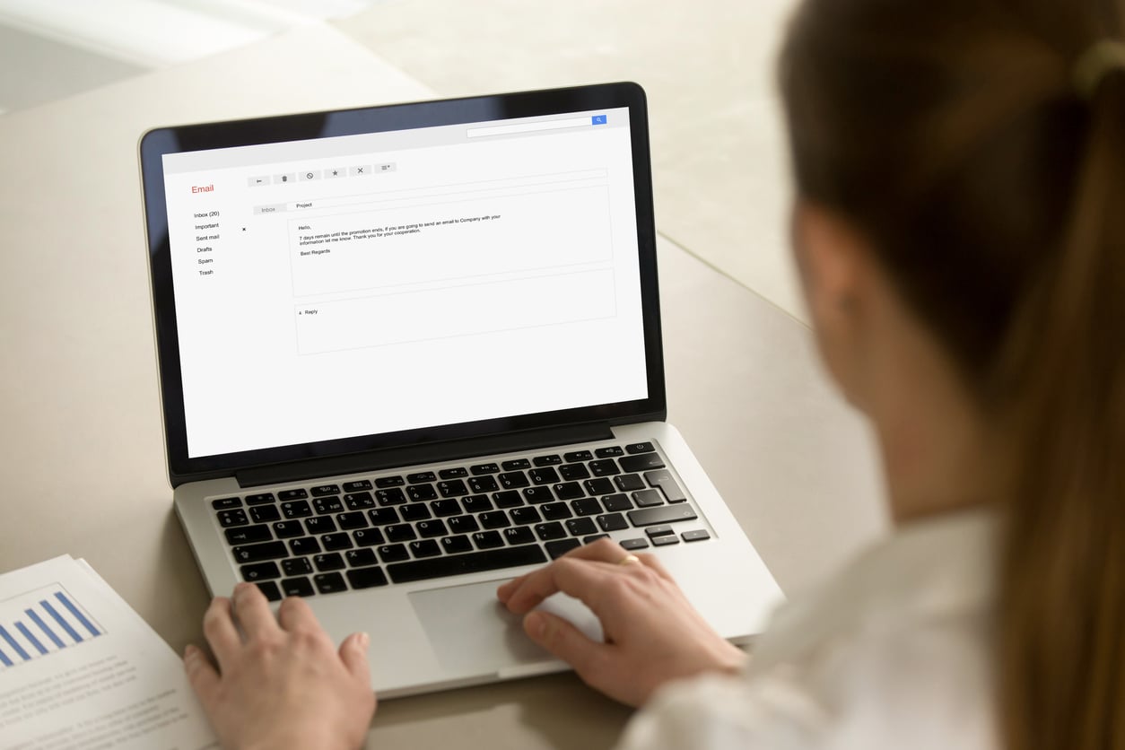 A woman is sitting at a desk using a laptop, composing an email about concert ticket hacks. Beside her, printed documents with blue charts are visible. The screen displays an email draft in a webmail interface.