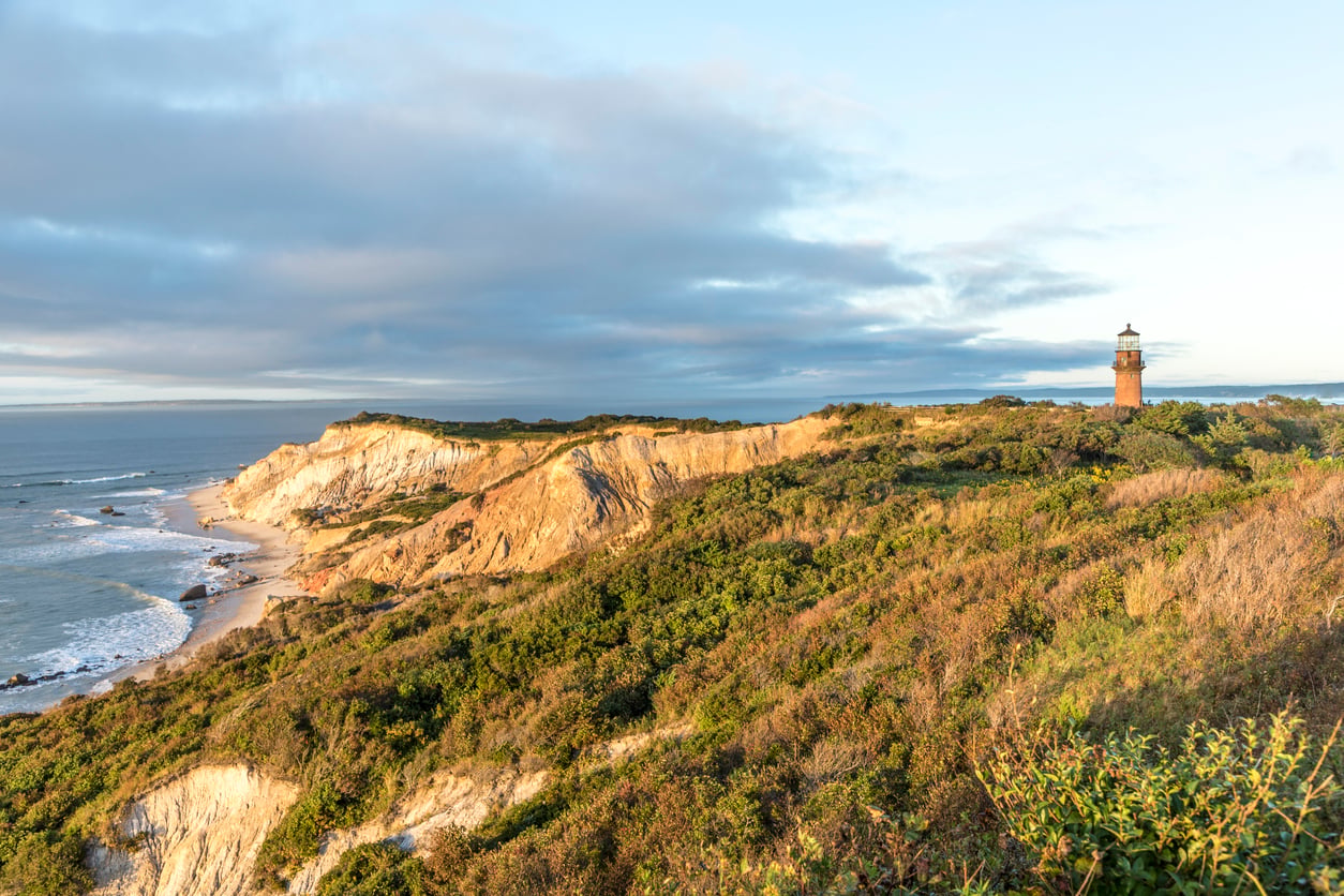 A lighthouse stands on a grassy cliff overlooking the ocean near charming beach towns, with dramatic rocky bluffs and waves crashing below under a partly cloudy sky at sunset.