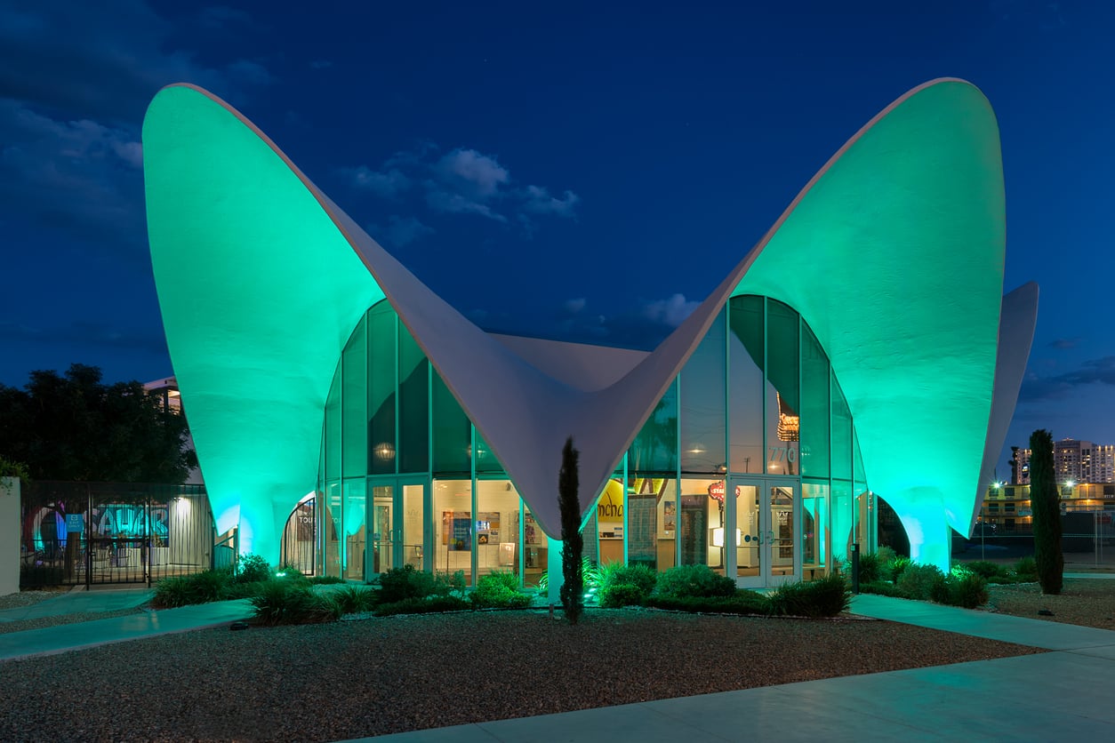 A modern building with sweeping, curved white rooflines and large glass windows is illuminated with green lights at night. For those seeking Las Vegas budget tips, the striking architecture can be admired for free as you stroll along the pathway lined with shrubs.