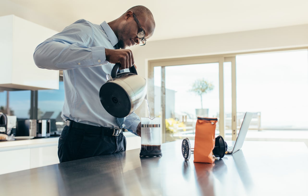 A man in business attire pours hot water from a kettle into a French press in a modern kitchen, enjoying one of his budget-friendly hobbies. A coffee bag, timer, and open laptop sit on the counter beside him.