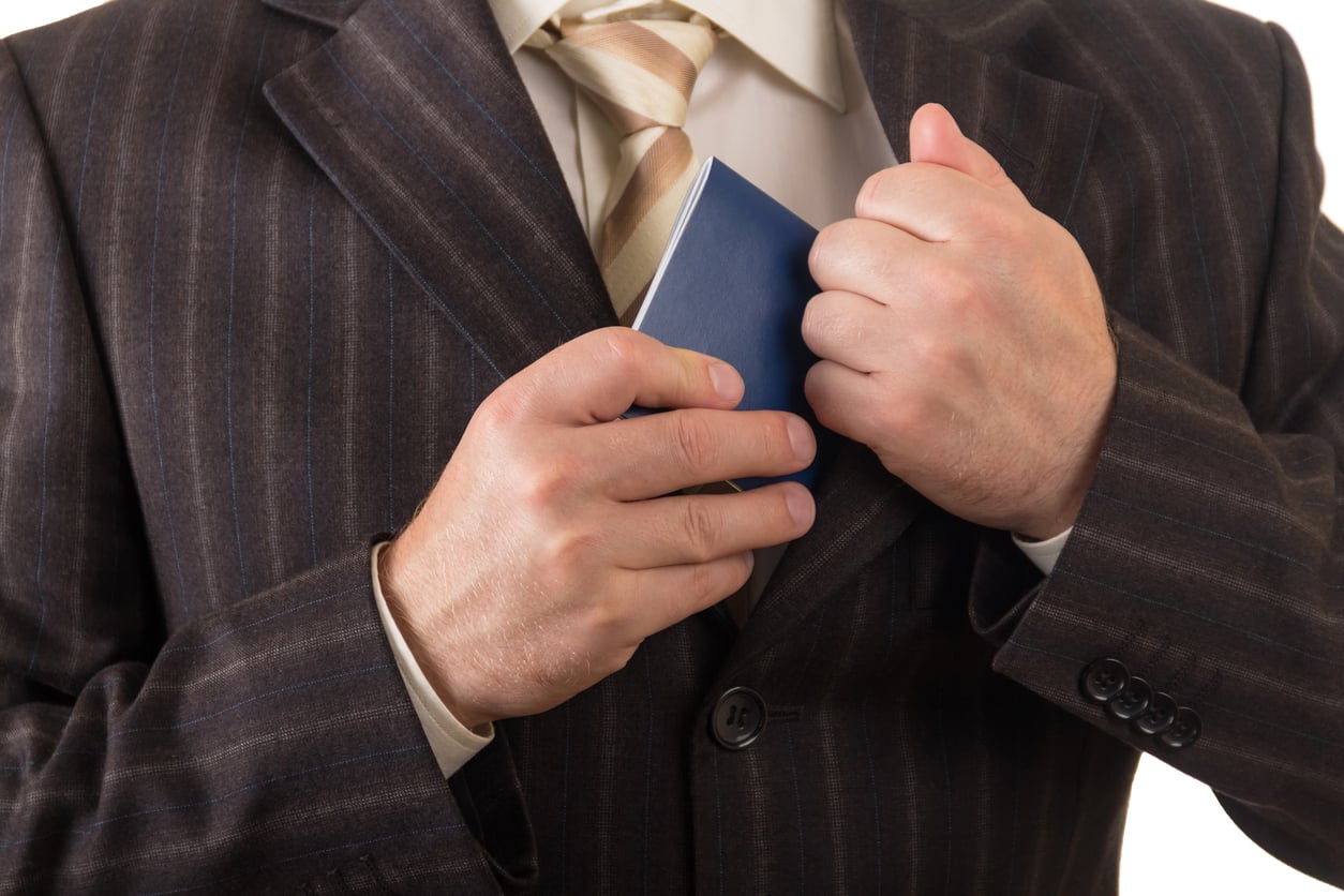 A man in a pinstripe suit and striped tie places a blue passport into the inside pocket of his jacket, practicing hotel room safety.
