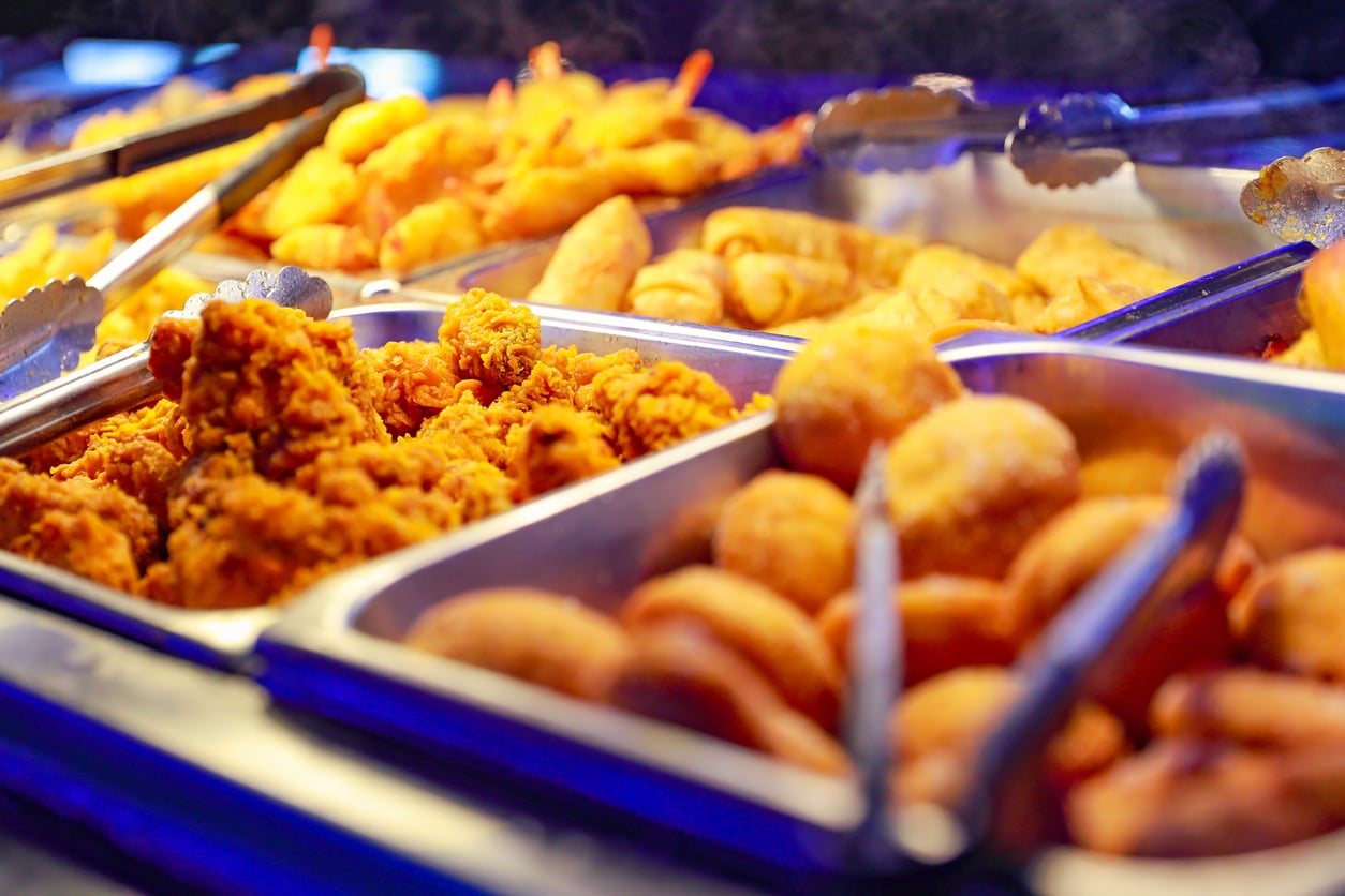 A buffet counter filled with various fried foods, including fried chicken, shrimp, egg rolls, and doughnuts, with metal tongs placed on the trays.