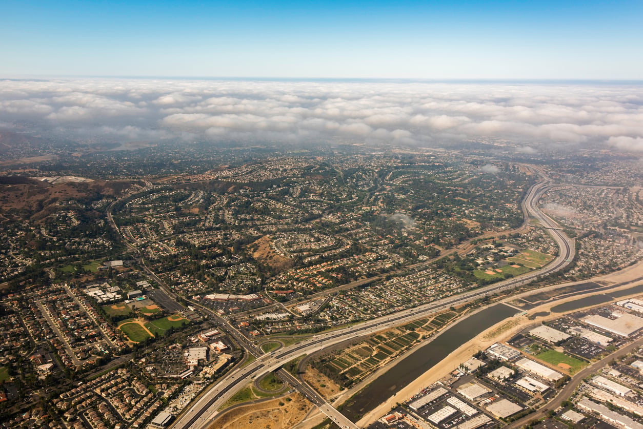 Trader Joe's New Store Openings. Aerial view of a sprawling suburban area with winding streets, highways, and a river. Scattered homes and green spaces are visible under a partially cloudy sky. The horizon shows low hills beneath a layer of clouds.