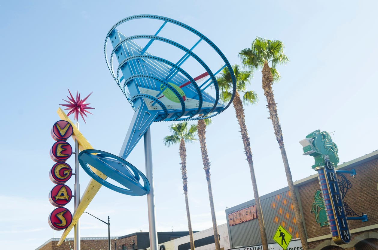 A large, blue martini glass sculpture stands upright next to a retro "VEGAS" neon sign and tall palm trees against a clear sky, offering a classic photo op for travelers seeking Las Vegas budget tips amid vibrant storefronts and signs.