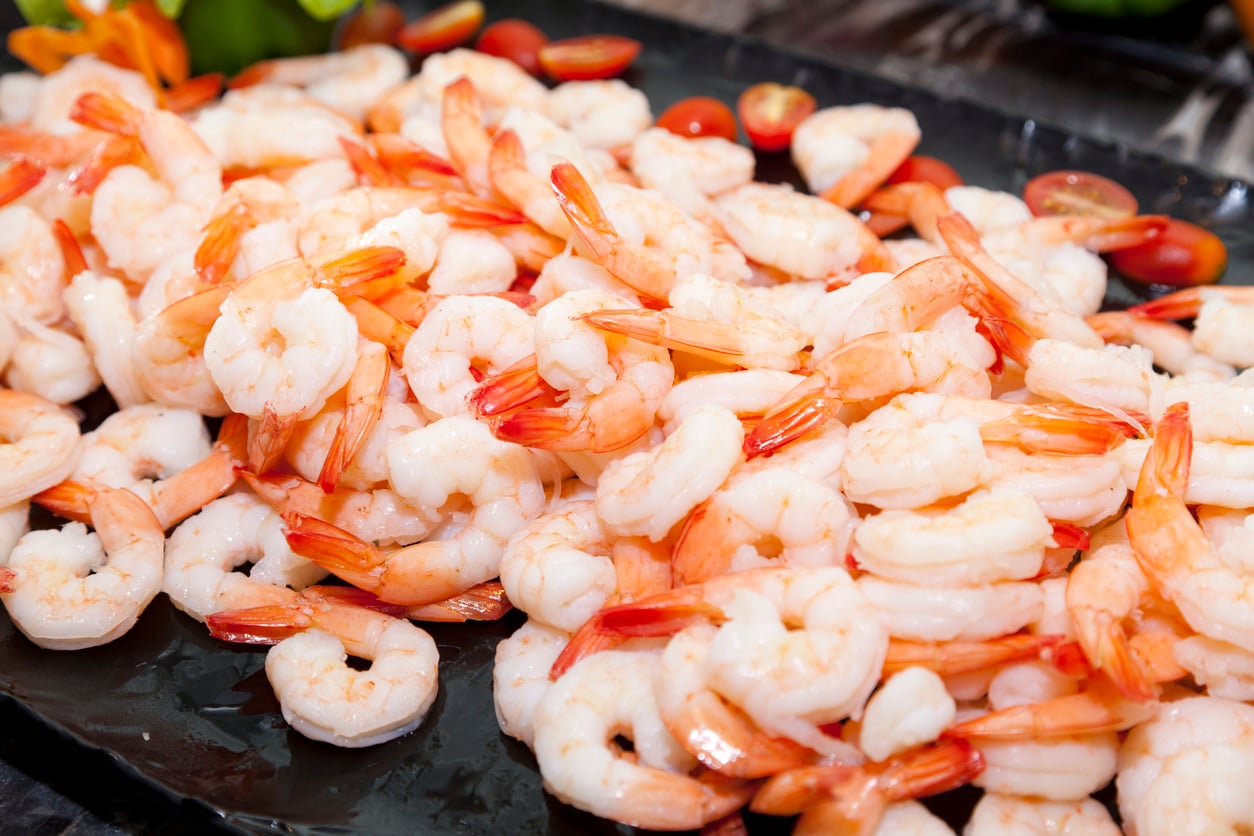 A close-up of a large pile of cooked shrimp with orange-tinted tails, displayed on a dark platter. Some cherry tomatoes are visible in the background.