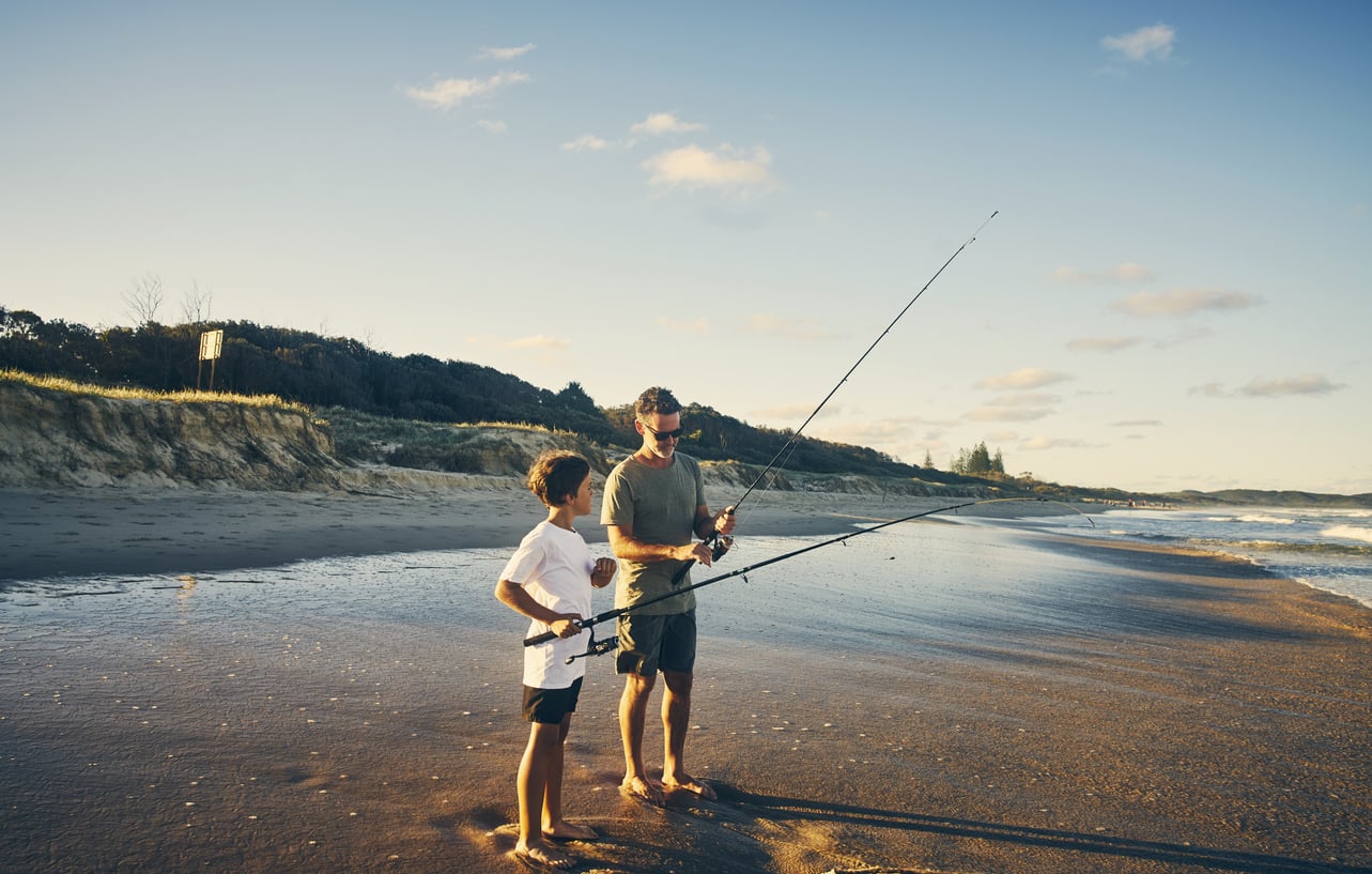 A man and a child stand on a sandy beach, enjoying budget-friendly hobbies as they fish together. The sun is low in the sky, casting warm light over gentle waves and grassy dunes in the background.