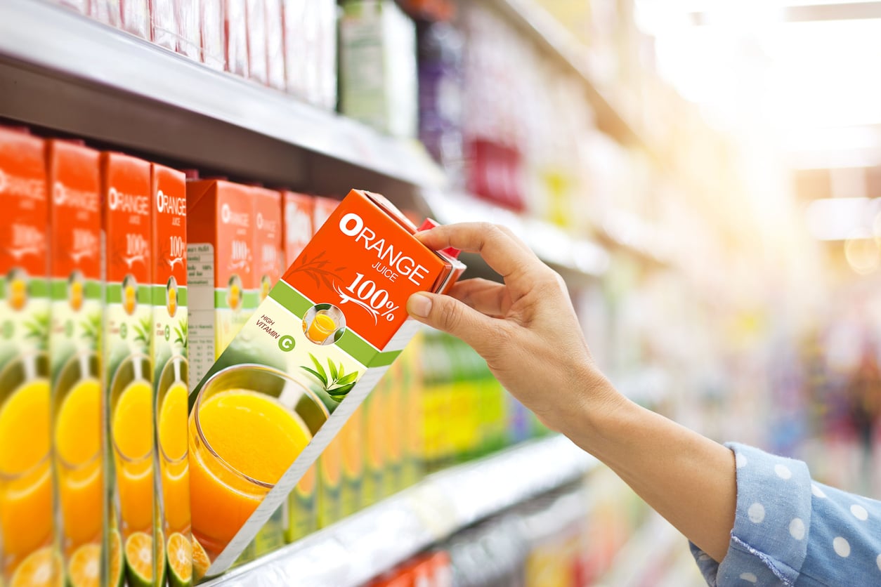 A person wearing a blue polka-dot shirt is picking up a carton of 100% orange juice from a supermarket shelf filled with similar, possibly overpriced products.