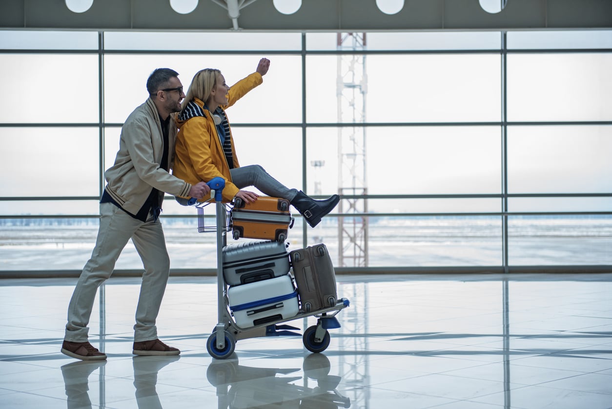 A man pushes a woman sitting on a luggage cart full of suitcases in an airport terminal. The woman is smiling and raising her arm as if waving or celebrating. Large windows show an outside view.