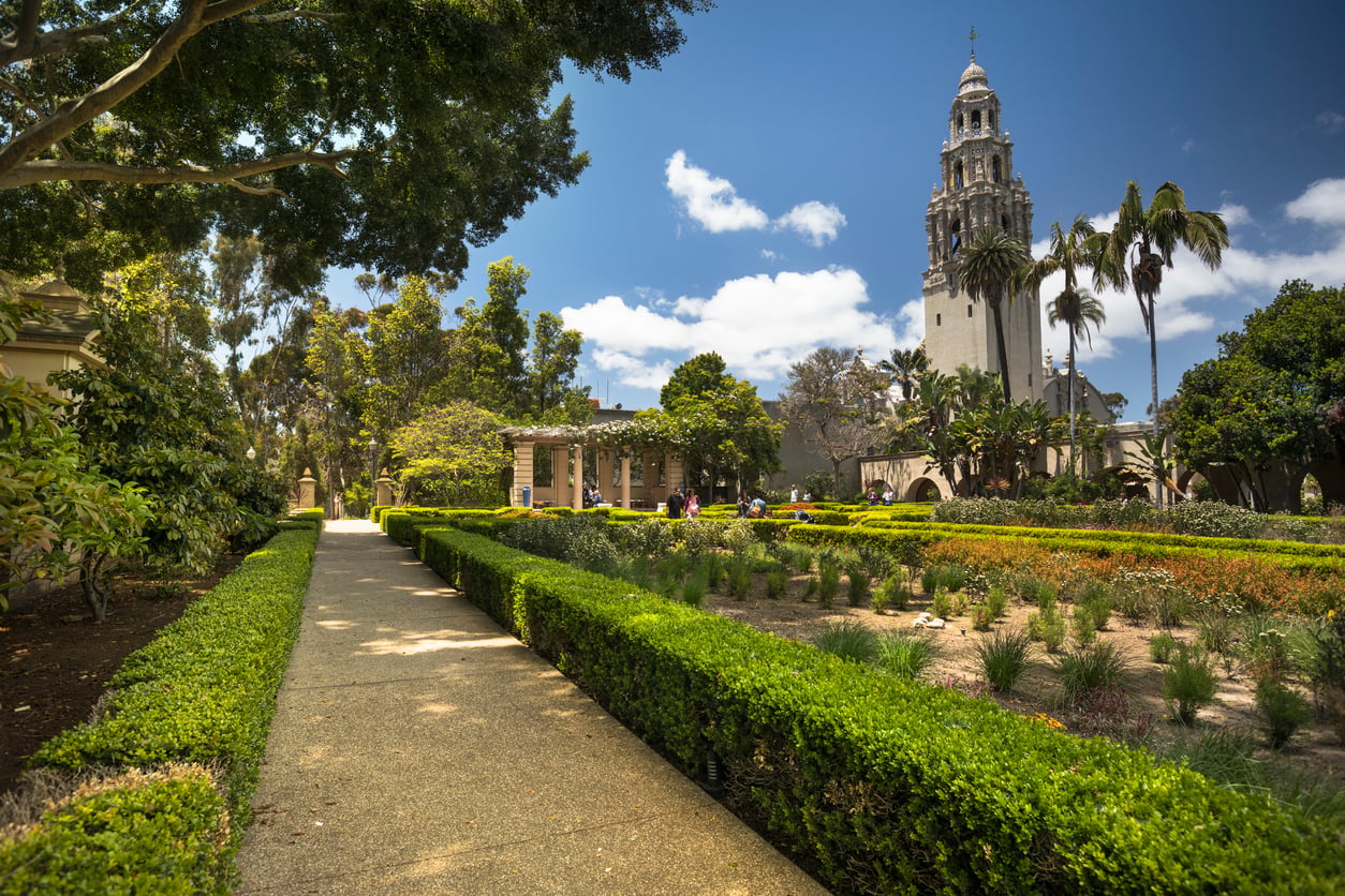 A paved garden path bordered by manicured hedges leads to a historic tower, offering Florida alternatives with palm trees and lush greenery under a blue sky with scattered clouds.