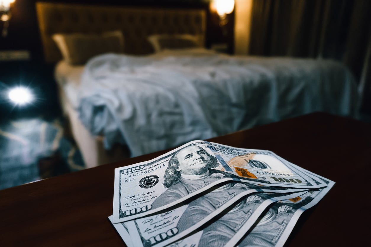 Three one-hundred dollar bills are spread out on a dark wooden table with an unmade bed and dim lighting in the background, hinting at Vegas money-saving tips for savvy travelers.