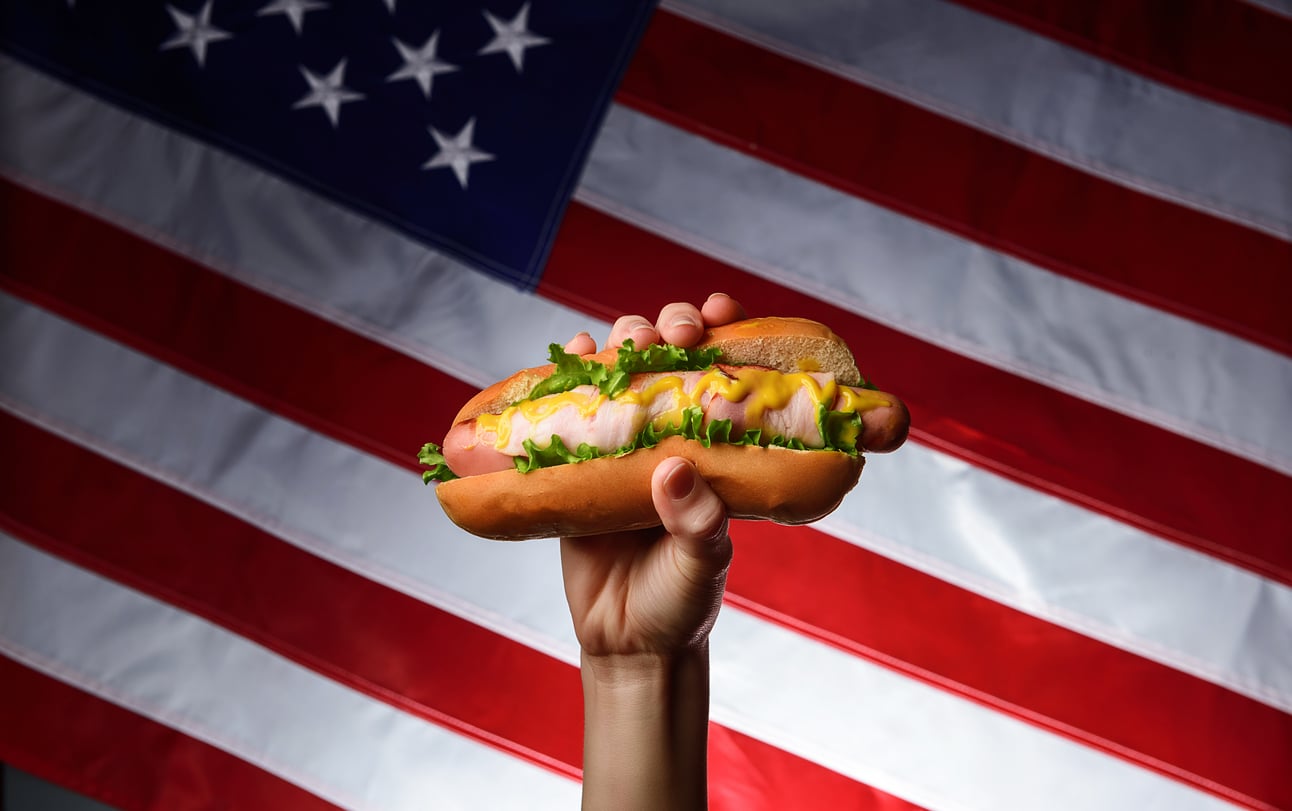 A hand holds up a classic fast-food hot dog with lettuce, mustard, and relish in front of a large American flag in the background.