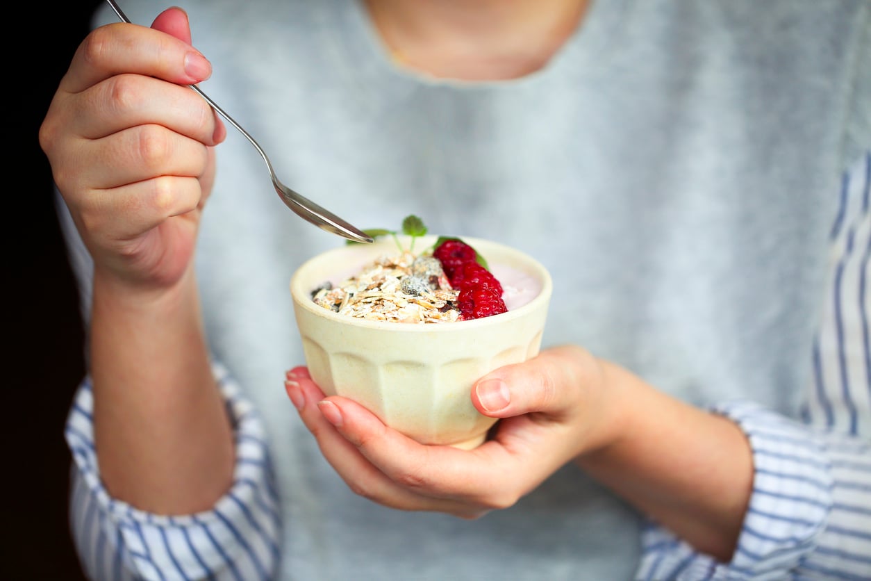 A person holds a bowl of yogurt topped with granola and berries in one hand and a spoon in the other, preparing to eat. The person, interested in egg substitutes, wears a light gray top with blue-and-white striped sleeves.