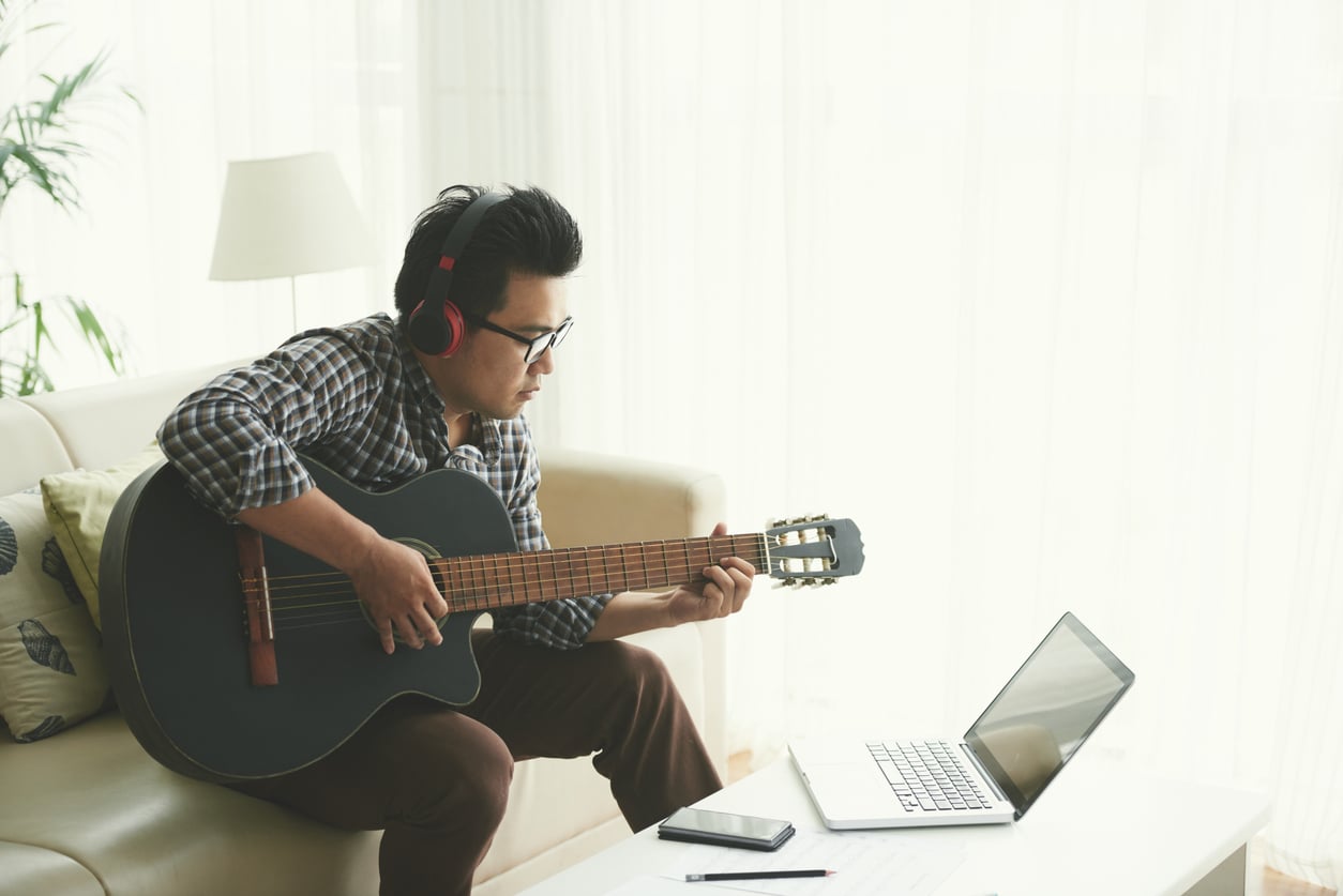 A man wearing headphones plays an acoustic guitar, enjoying budget-friendly hobbies while sitting on a couch and looking at a laptop on a coffee table in a bright living room. A smartphone and notepad are also on the table.