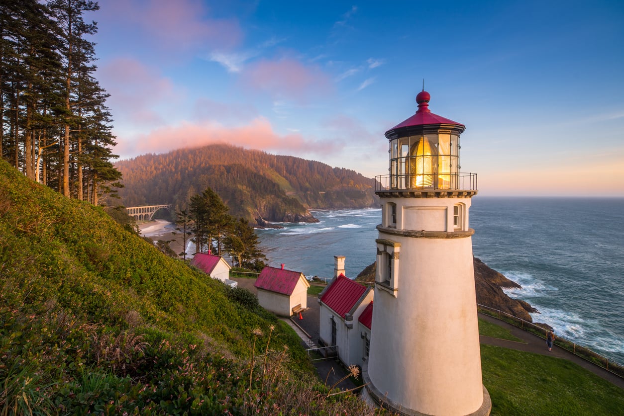 A white lighthouse with a red roof stands on a green cliff overlooking the ocean at sunset, with nearby buildings, a forested hillside, and a distant arched bridge under a colorful sky.