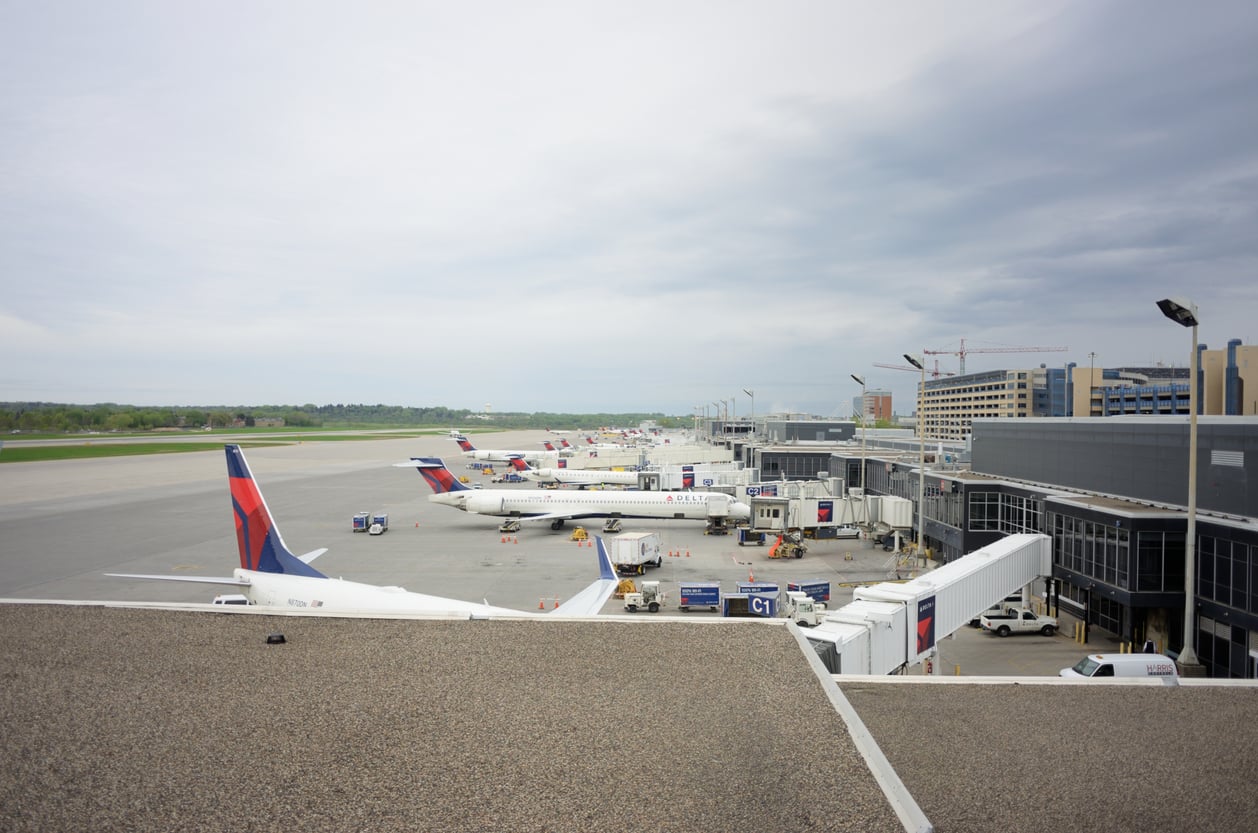 Several airplanes are parked at gates at a convenient airport terminal under a cloudy sky, with jet bridges connecting to the building and ground vehicles nearby. A runway and taxiway stretch into the distance.