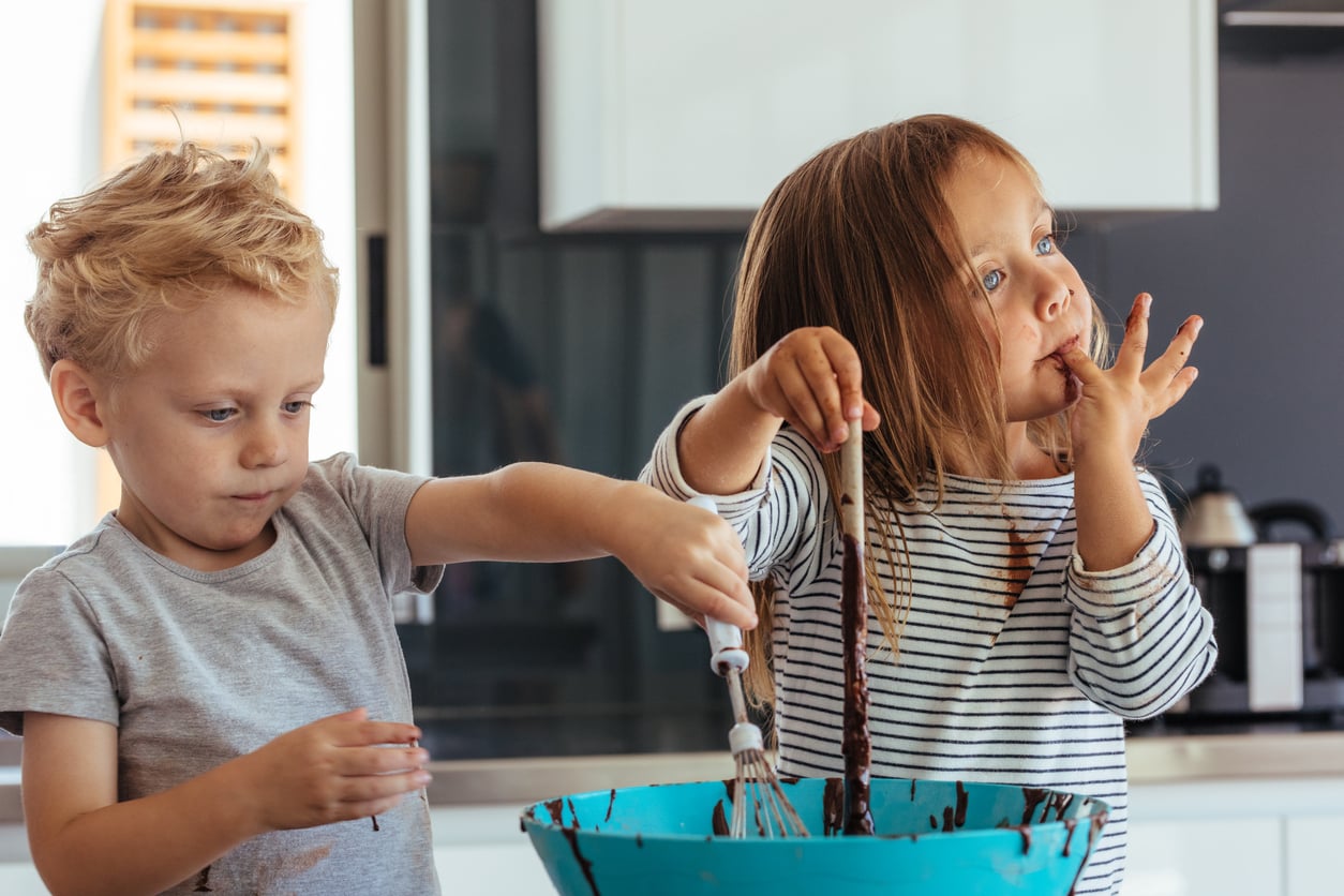 Two young children stand in a kitchen mixing chocolate batter in a blue bowl. The scene, familiar to many middle class moms, shows the girl licking chocolate off her finger while the boy mixes with a spoon, both focused on baking.