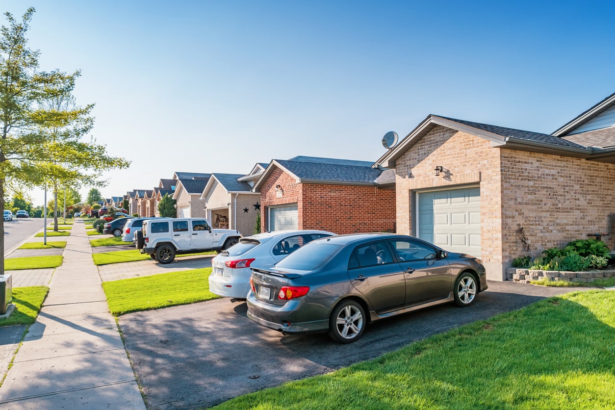 A row of modern suburban houses with brick exteriors, each with a driveway and cars parked outside—some showing signs of delayed purchases—under a clear blue sky on a sunny day.