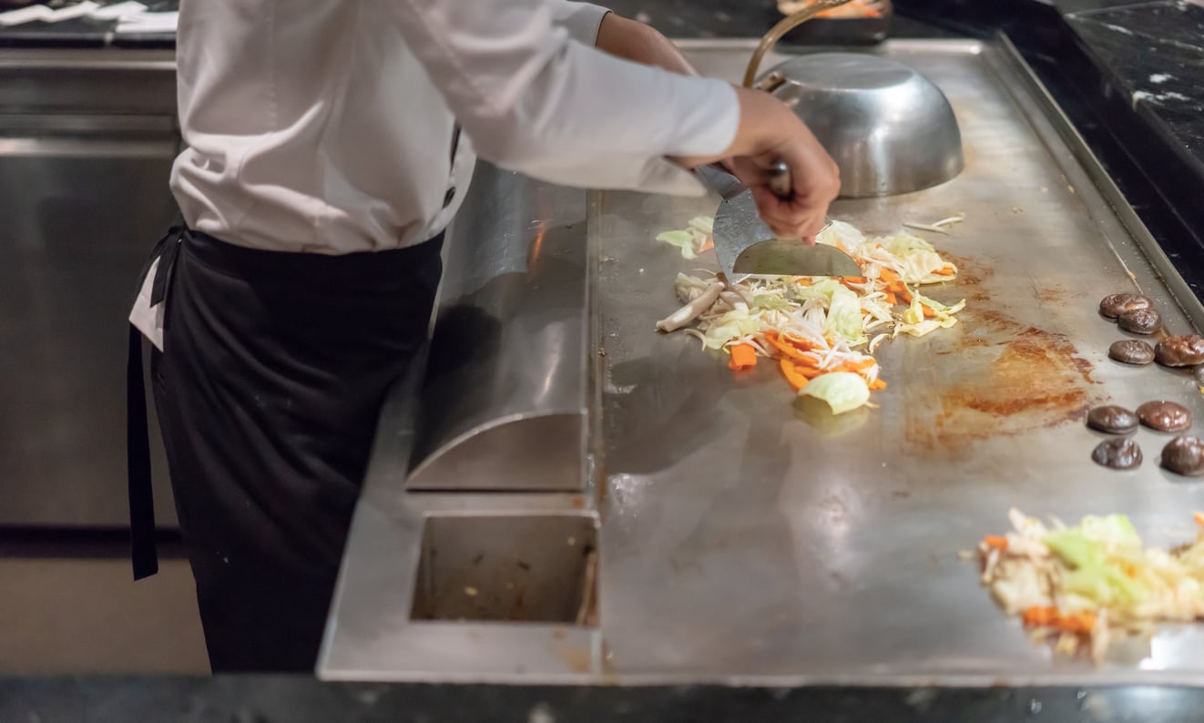 A chef in a white shirt and black apron cooks sliced vegetables on a teppanyaki grill, using a spatula to stir-fry cabbage, carrots, and mushrooms.