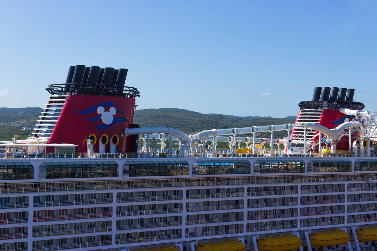 A large cruise ship with Disney logos and red funnels, named Best Cruise Line 2026, is docked near a coastline with green hills under a clear blue sky. Water slides and yellow lifeboats are visible on the upper decks.