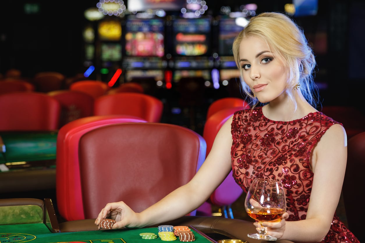 A woman in a red, sequined dress sits at a casino table holding a glass of brandy and poker chips, with slot machines and red chairs in the background—capturing the thrill before exploring Vegas money-saving tips.