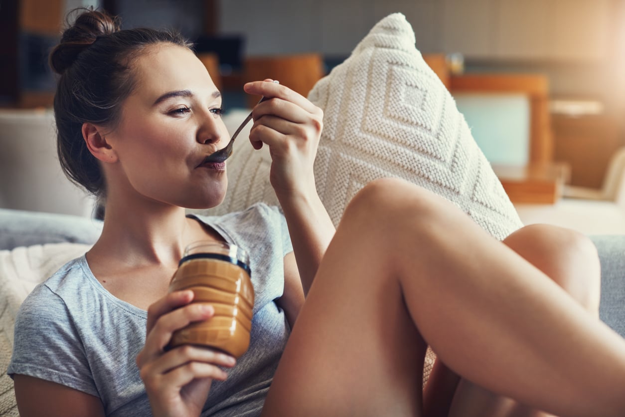 A woman sits on a couch with her legs up, holding a jar from one of the best peanut butter brands and eating from it with a spoon, looking content and relaxed.