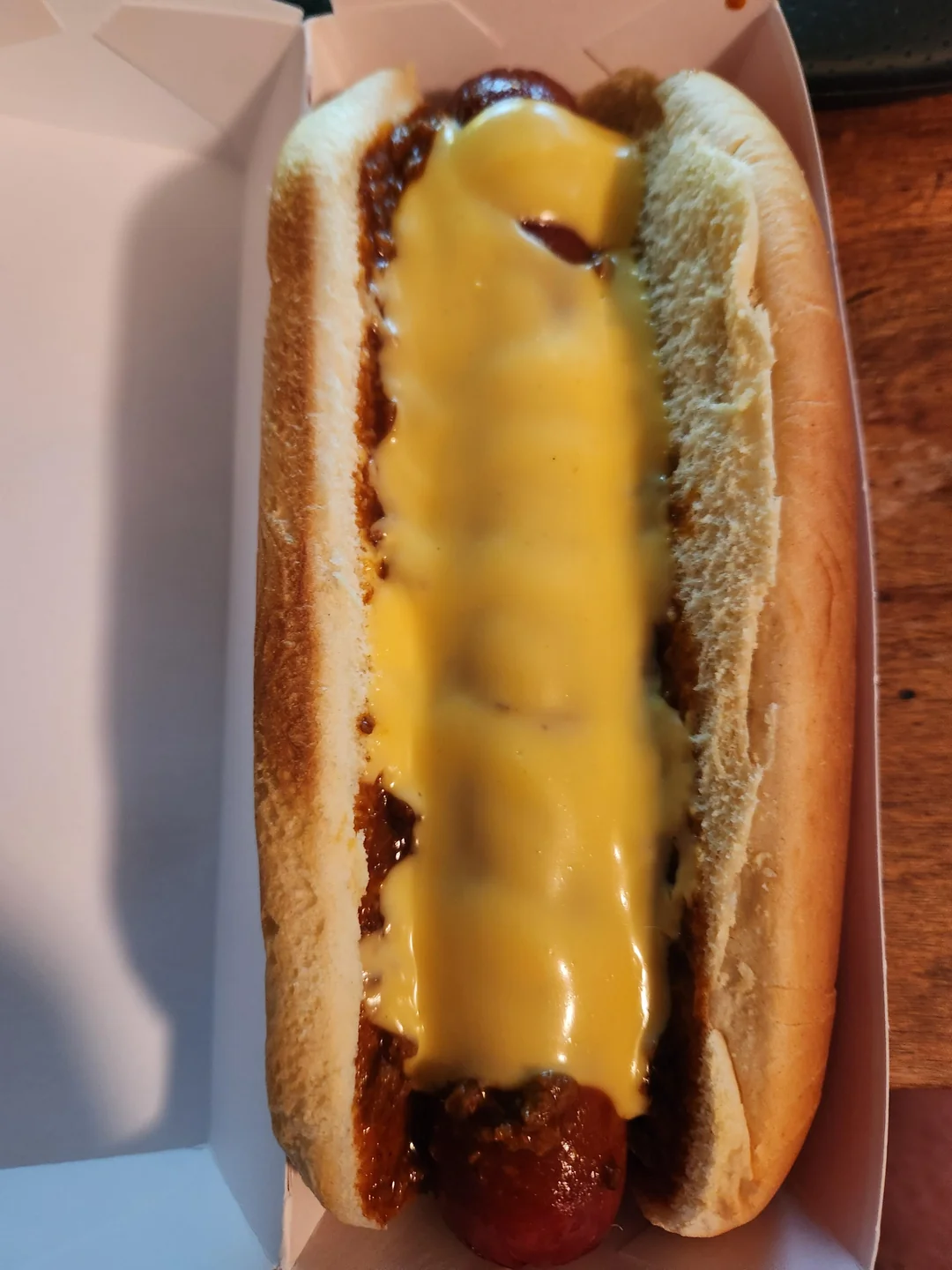 A close-up of a Fast-Food Hot Dog in a bun, topped with chili and melted cheese, served in a white paper tray on a wooden surface.
