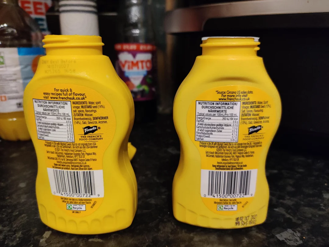 Two yellow squeeze bottles of French's mustard are placed side by side on a kitchen counter with their nutrition labels facing the camera. Other bottles and kitchen items are visible in the background.