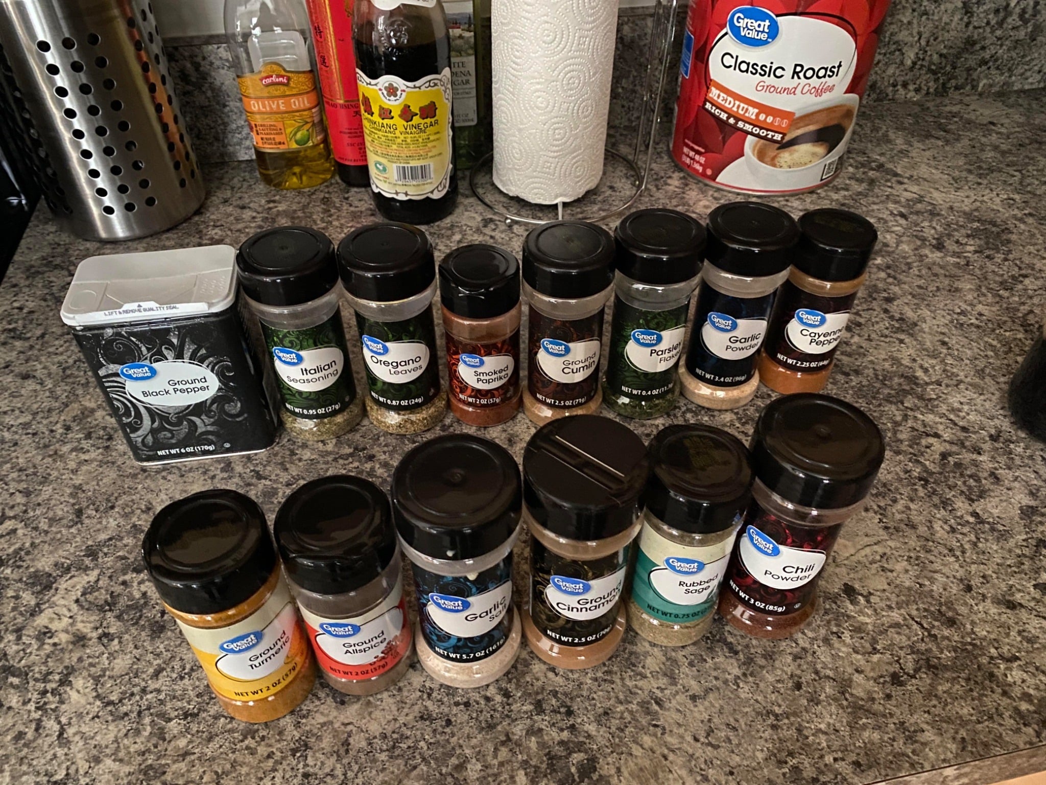 A variety of spice and seasoning containers, including Walmart products like Great Value, are arranged on a kitchen countertop. Visible items include ground black pepper, garlic powder, and chili powder, with other kitchen essentials in the background.