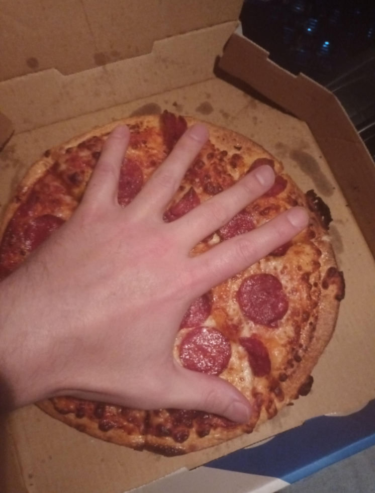 A hand with fingers spread is placed on top of a pepperoni pizza inside an open cardboard box, showing the size comparison between the hand and the pizza.