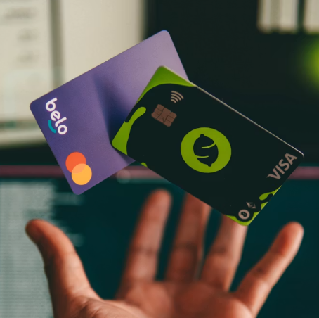 A hand is holding two credit or debit cards—a purple Belo Mastercard and a black Visa card with green accents—against a blurred computer screen, showing that you don't have to pay more for less when choosing your payment options.
