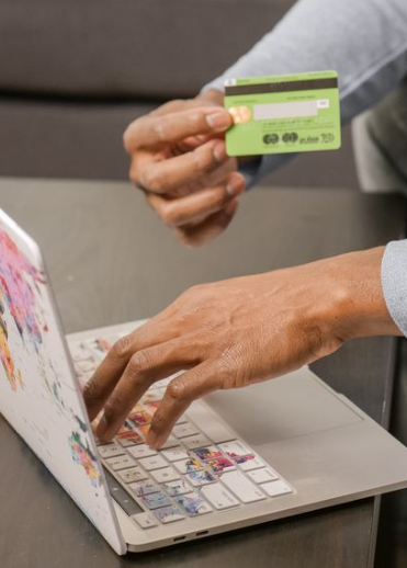 A person holding a green credit card in one hand while typing on a colorful, paint-splattered laptop keyboard with the other, unaware they're about to pay more for less.