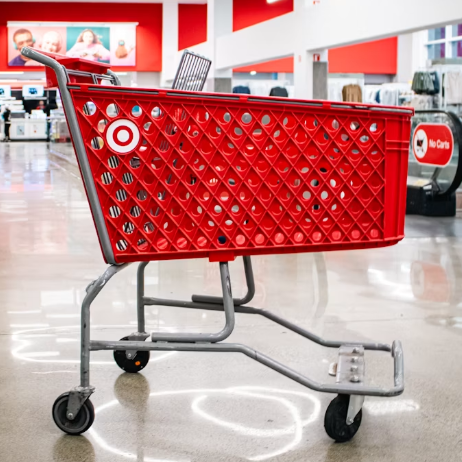 A red Target shopping cart stands empty inside a brightly lit Target store, where shelves and checkout counters await shoppers ready to pay more for less.