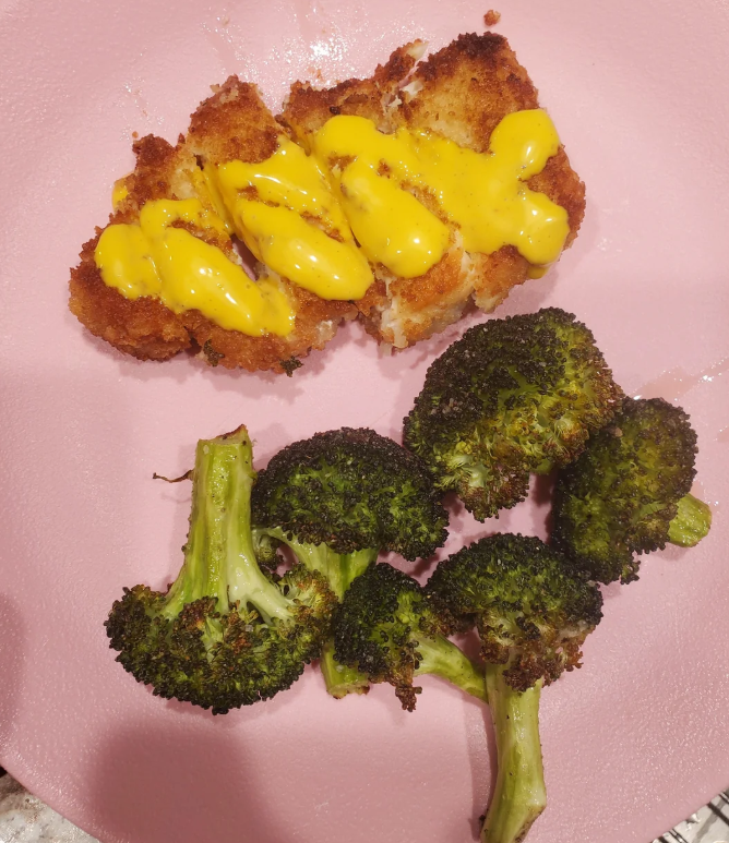 A pink plate with breaded fish topped with yellow cheese sauce and a serving of roasted broccoli florets—making you wonder if you're starting to pay more for less at your favorite spot.