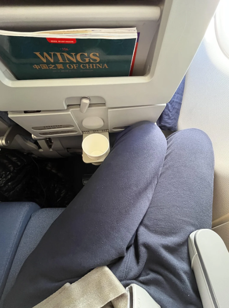 A person sitting in an airplane seat with a tray table up, a paper cup in the holder, and a "Wings of China" magazine in the seat pocket—showcasing how travelers now often pay more for less comfort, wearing dark pants and a seatbelt.