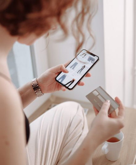 A person holding a smartphone displaying online clothing shopping and a credit card, sitting by a window with a cup of tea or coffee on the floor nearby, wonders if they’ll pay more for less with each purchase.